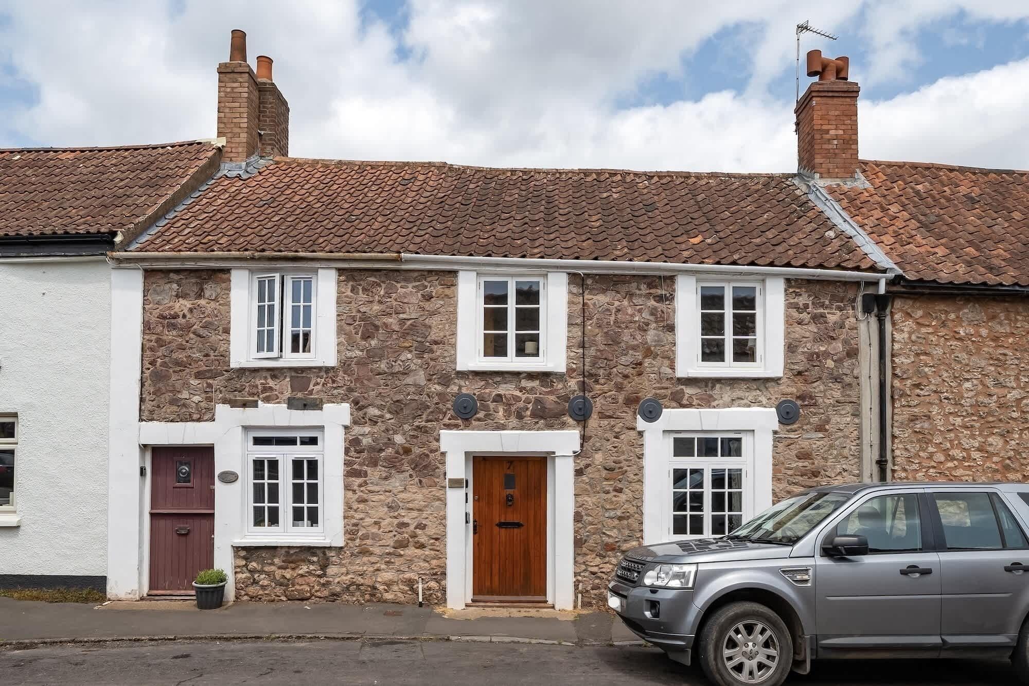Une maison en pierre à deux étages, avec un toit recouvert de tuiles rouges, des fenêtres à cadre blanc et un garage pouvant accueillir une seule voiture.