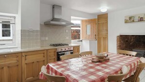 A kitchen with a gas stove, wooden cabinets, a tiled backsplash, and a checkered tablecloth-covered dining table. - Historic Cottage & Garden nr Quantock Hills (Nether Stowey)