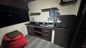 Une cuisine compacte équipée d’un réfrigérateur, d’un distributeur d’eau et d’une cuisinière. On y trouve également un petit tabouret et une fenêtre donnant sur l’extérieur.