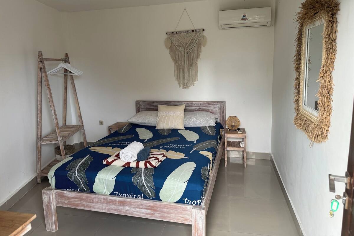 A bedroom with a bed, a wooden ladder, a macrame wall hanging, a mirror, and a small table with a lamp.