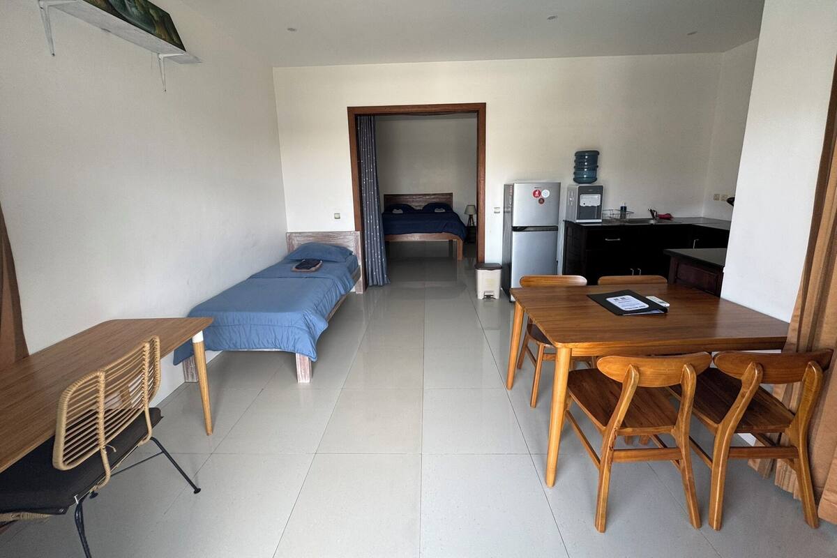 A compact room with a bed, a small desk, a chair, a table, and a kitchenette.