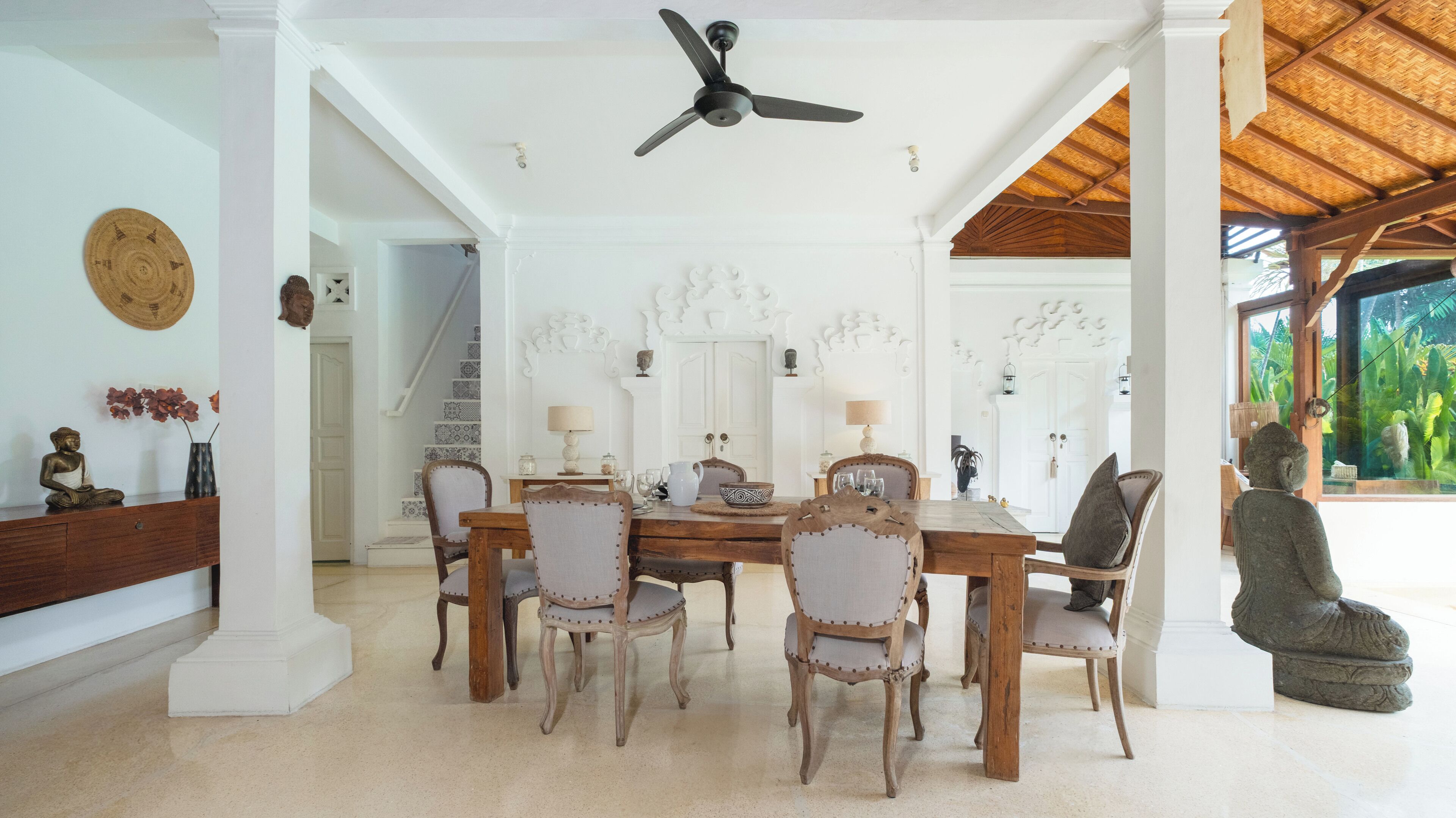 A dining area with a wooden table and chairs, a ceiling fan, and a view of a tropical garden.