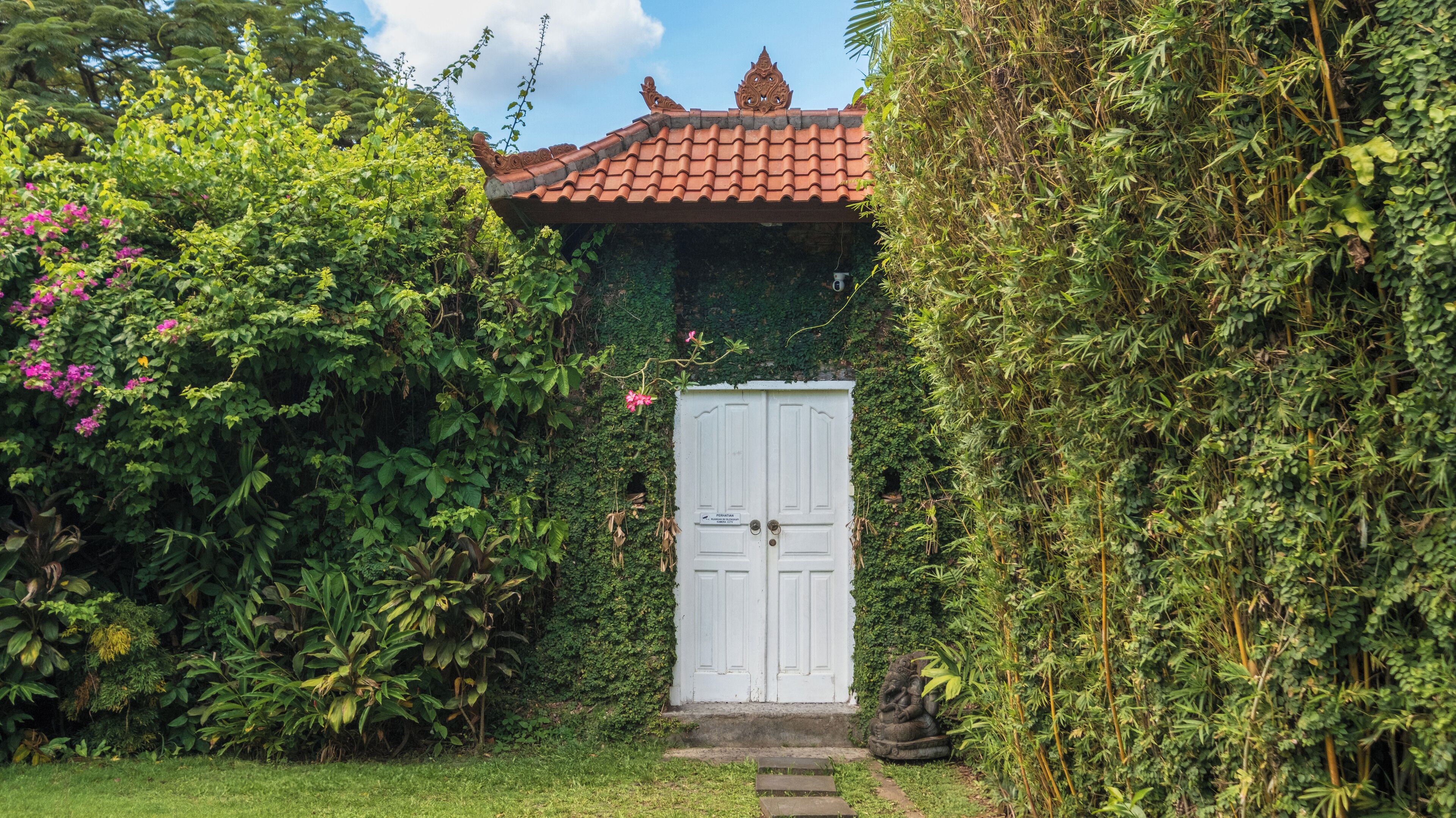 A white door with a sign, surrounded by lush greenery and a tiled roof.