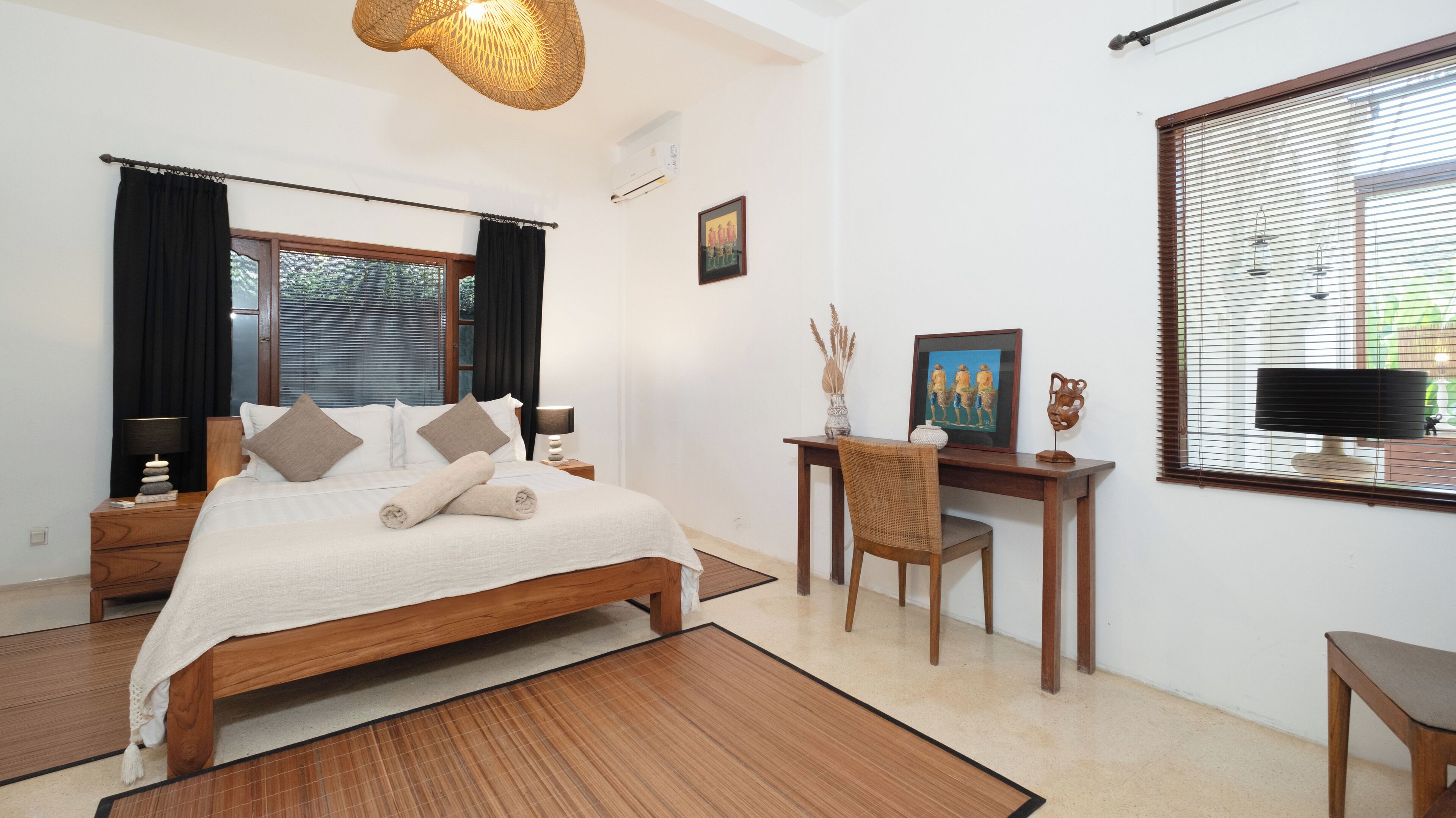 A bedroom with a wooden bed, a desk, and a chair. There is a window with blinds, a wall-mounted air conditioner, and a painting on the wall.