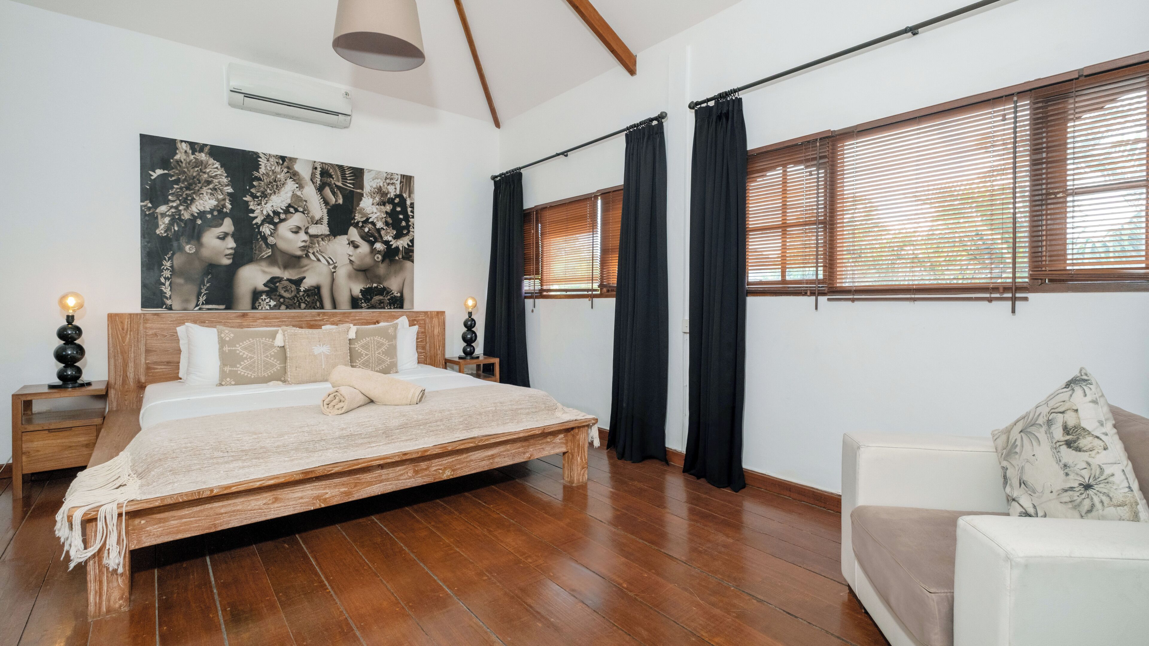 A spacious bedroom with a large bed, a wooden headboard, a bedside table with a lamp, a wall-mounted air conditioner, and a large window with blinds.