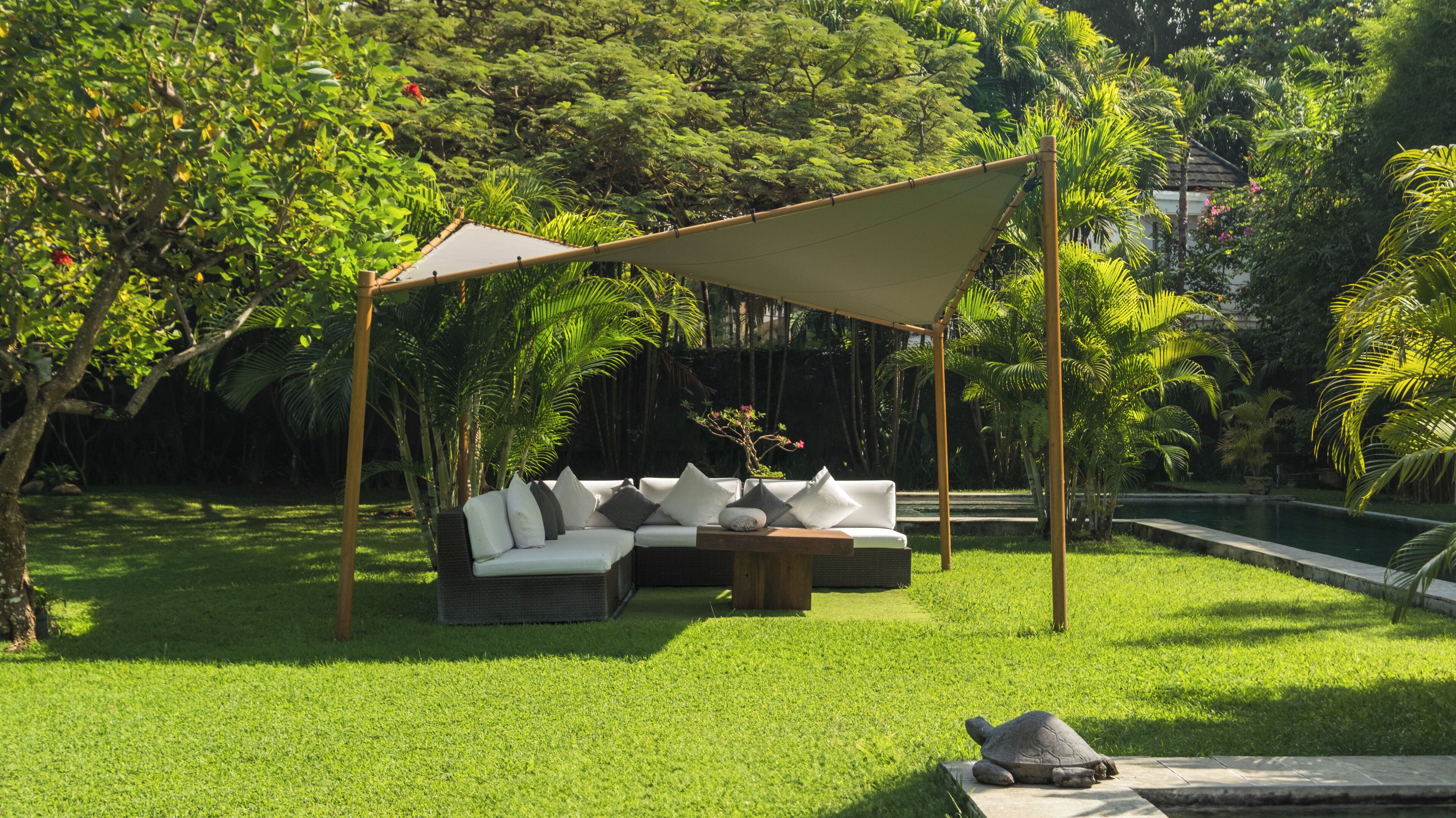 An outdoor seating area with a sofa and table under a shaded canopy, surrounded by lush greenery and a small pond.
