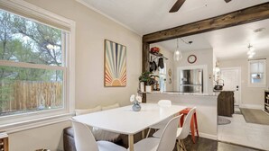 Een eethoek met een witte tafel en stoelen, een houten plafond en uitzicht op de achtertuin door een raam.