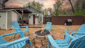 Een achtertuin met een vuurkuil, omringd door blauwe Adirondack-stoelen, een overdekte patio en een barbecue.