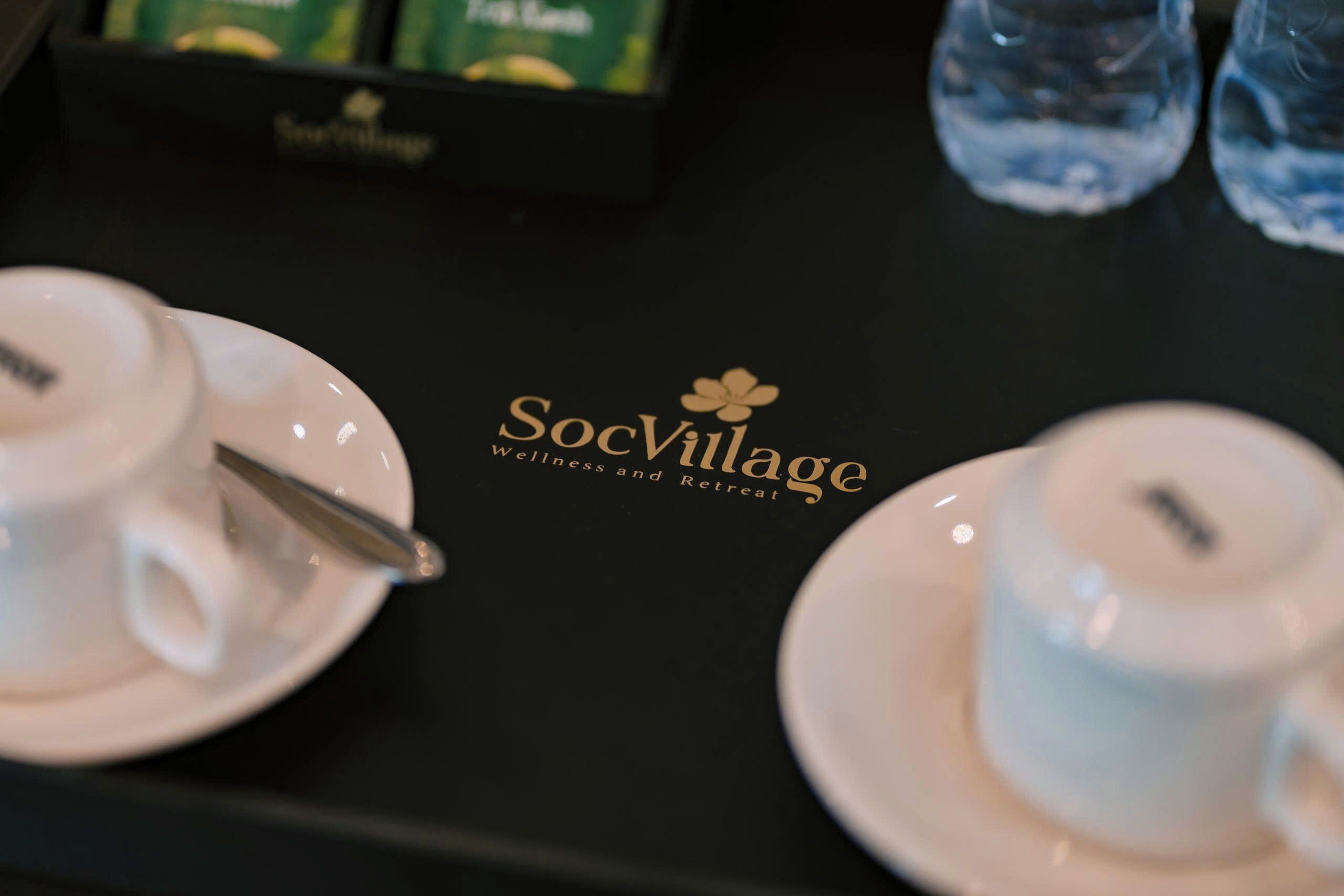 A table with two white cups and saucers, a spoon, and a box labeled "SocVillage Wellness and Retreat".