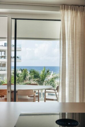 A room with a large window overlooking a balcony with a view of the sea and palm trees.
