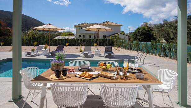 A poolside dining area with a wooden table, white chairs, and a view of a house in the background.