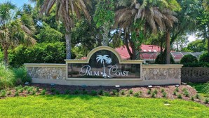 A sign for Palm Coast with a palm tree logo, surrounded by lush greenery and a stone wall. - Market View Condo | European Village | Minutes to Beach & Restaurants (Palm Coast)
