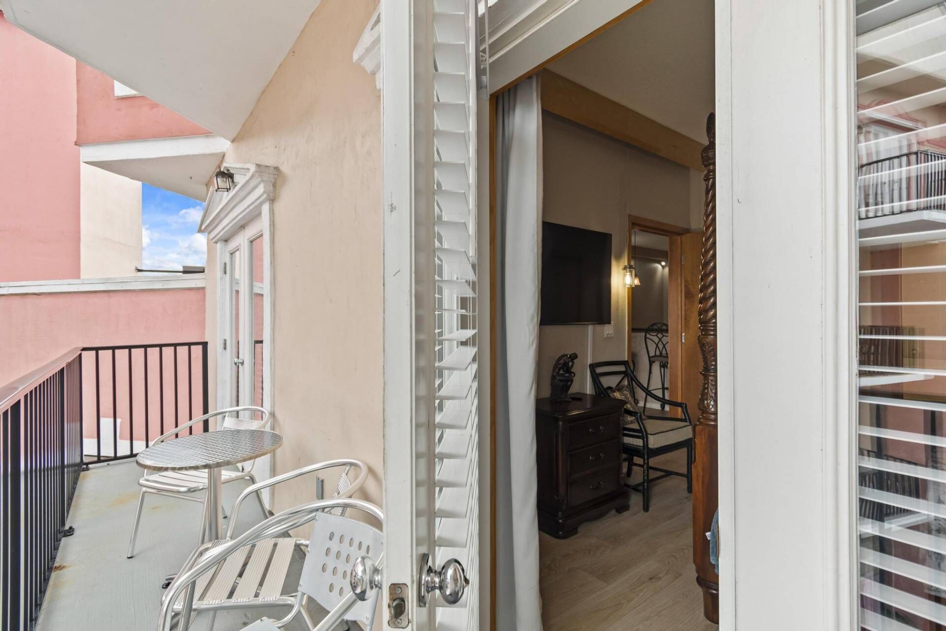 A balcony with metal chairs and a small table, a wooden chair inside, and a bookshelf with a TV on the wall.