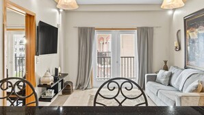 Ein Wohnzimmer mit einer Couch, einem Esstisch mit Stühlen, einem an der Wand montierten Fernseher und einem Balkon mit Vorhängen.