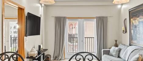 A living room with a sofa, a dining table with chairs, a TV mounted on the wall, and a balcony with curtains.