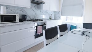A modern kitchen with white cabinets, a marble backsplash, a gas stove, a microwave, and a dining area with a glass table and black chairs. - 2-bed Gucci Flat - Sleeps 6 & Close to Harrods (London)