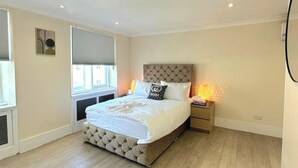 A bedroom with a large bed, a headboard, bedside tables, a window with blinds, and a radiator. - 2-bed Gucci Flat - Sleeps 6 & Close to Harrods (London)