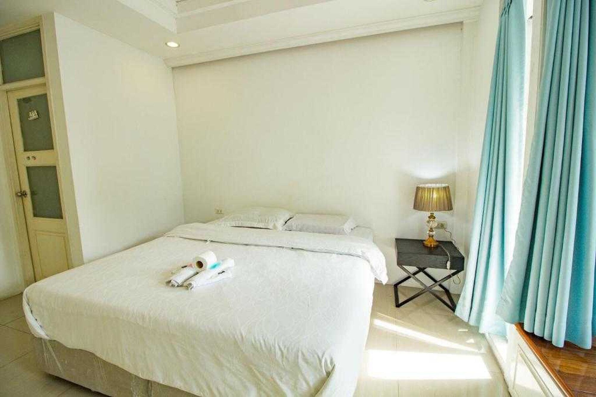 A hotel room with a bed, a bedside table with a lamp, a window with curtains, and a door with frosted glass panels.