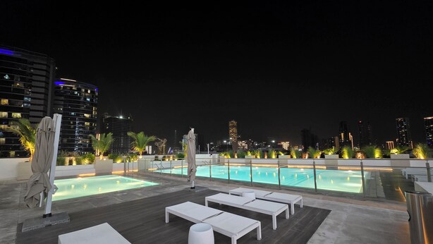 A rooftop pool area with lounge chairs and umbrellas, surrounded by city buildings at night. - 16th floor wonderland- 2 Bedroom (Dubai)
