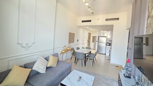 Una sala moderna con un sofá gris, una mesa de centro de mármol y una zona de comedor con mesa y sillas. Se observa una cocina al fondo con un refrigerador y una estufa.