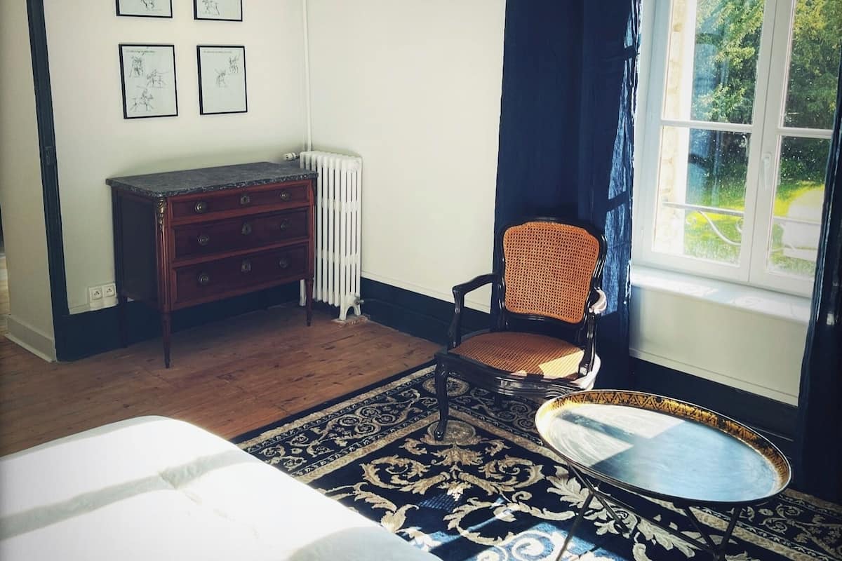 Une pièce comprenant un lit, une commode en bois, une chaise, une petite table et une fenêtre donnant sur des arbres.