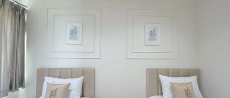 Habitación de hotel con dos camas individuales, cada una con cabecero color beige y ropa de cama blanca. Encima de cada cama hay cuadros enmarcados en la pared.