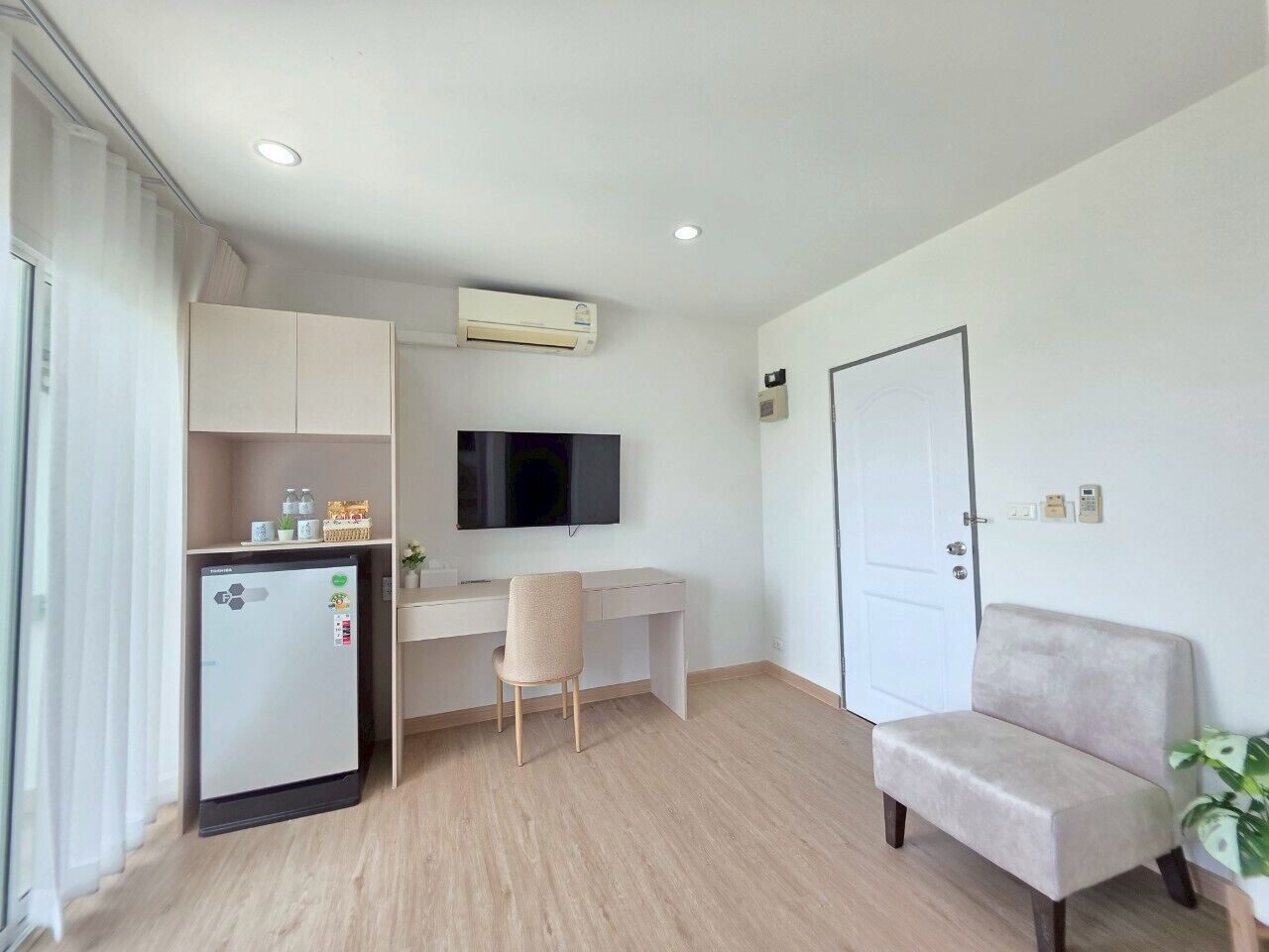 A modern, minimalistic room with a white desk, a chair, a sofa, a small kitchenette with a refrigerator, and an air conditioning unit.