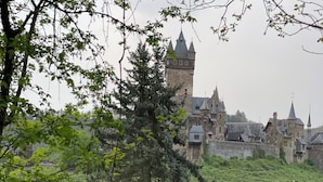 A medieval castle with a prominent tower, surrounded by lush greenery and a small building in the foreground. - Vacation apartment Cochem Mosel Pinnerbergstube (Cochem)