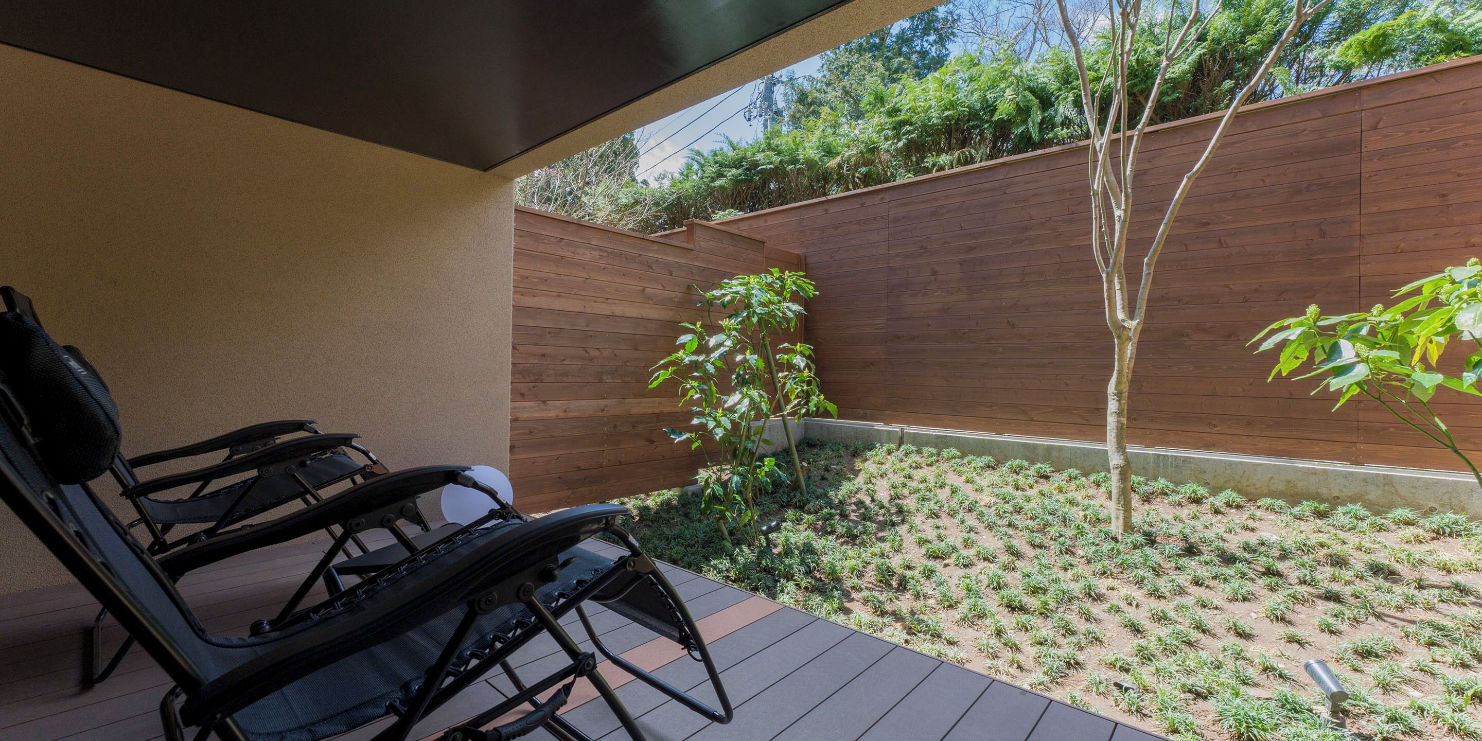 A patio area with a wooden deck, a black folding chair, and a small garden with plants and a tree.