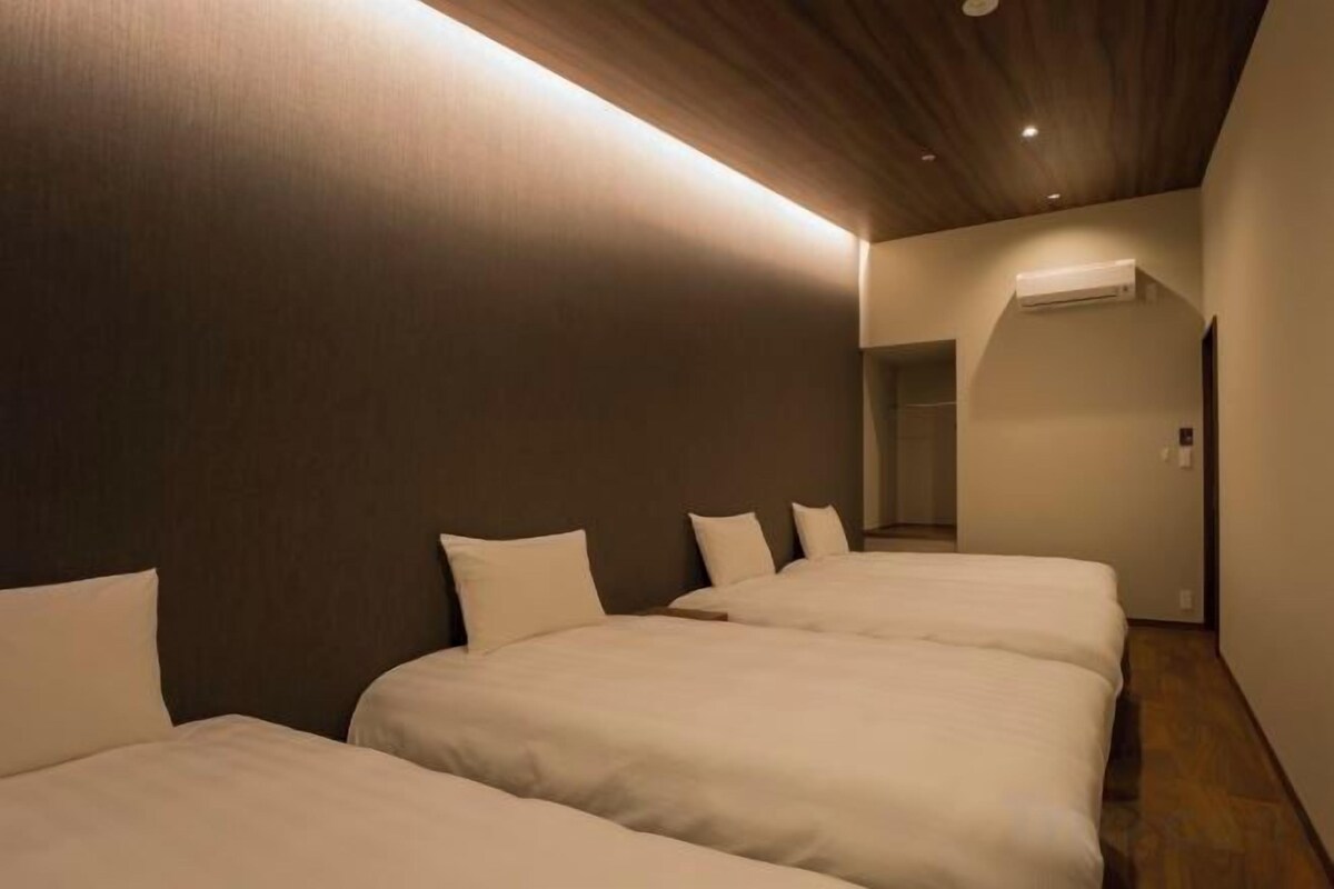 Une chambre d’hôtel avec trois lits alignés, un plafond en bois et un couloir menant à une porte.