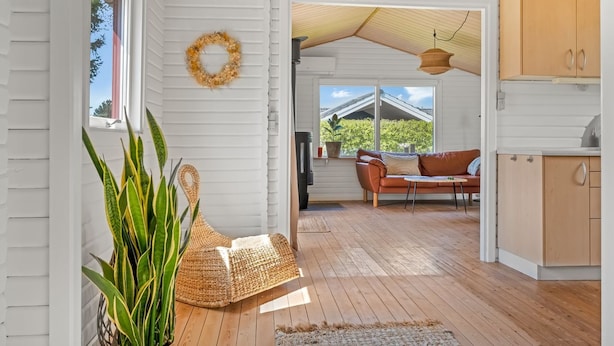 Un intérieur chaleureux avec un plafond en bois, un banc en osier, une plante en pot, un pouf en osier et une vue sur l’extérieur par une fenêtre.