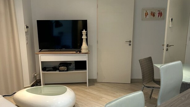 Ein modernes Hotelzimmer mit einem Flachbildfernseher auf einem Holzständer, einer runden Sitzecke, einem Esstisch und einer Tür mit dekorativer Verkleidung.