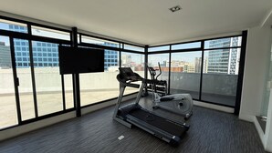 Un gimnasio moderno con cinta de correr, elíptica y pesas, amplias ventanas con vistas a la ciudad.