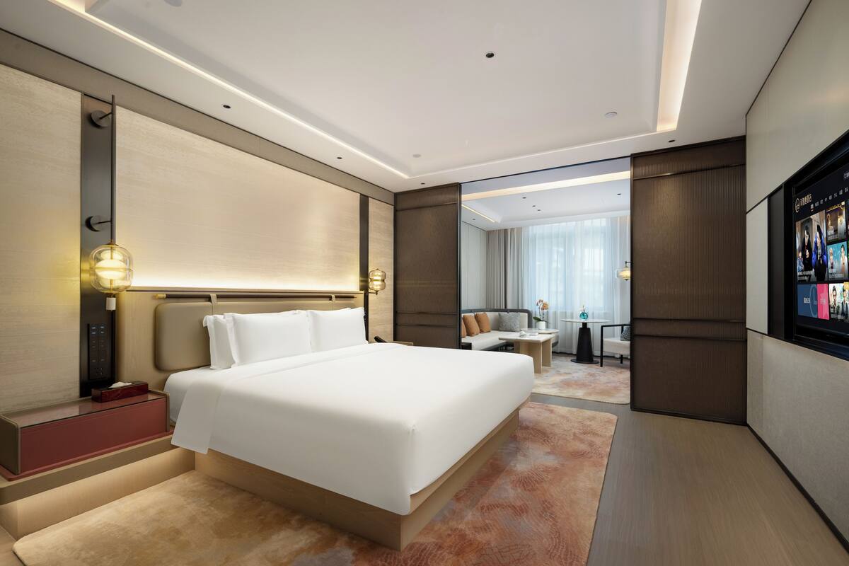 A modern hotel room with a large bed, bedside tables, a TV, and a view of the hotel lobby.