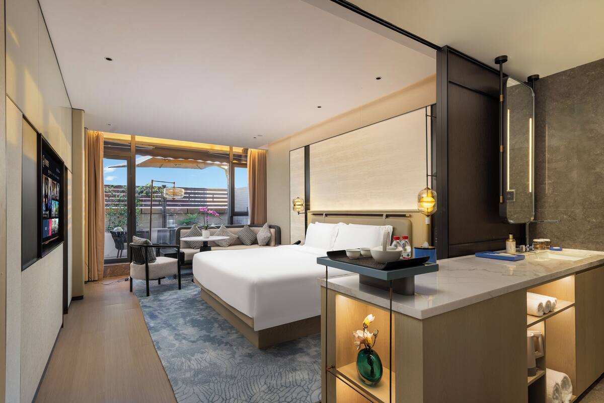 A modern hotel room with a large bed, a sitting area with a sofa and chairs, a small table, a bathroom with a mirror, and a view of the outdoors.