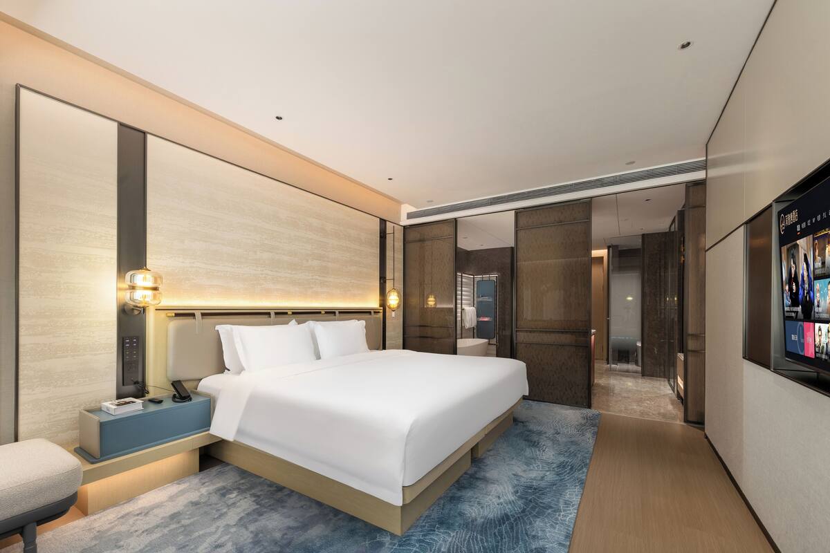 A modern hotel room with a large bed, bedside tables, a TV, and a bathroom visible through a glass door.