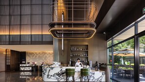 Un hall d’hôtel moderne avec un comptoir de réception en marbre, des tabourets de bar verts, une grande fenêtre donnant sur l’extérieur et un espace bar avec des étagères remplies de bouteilles.