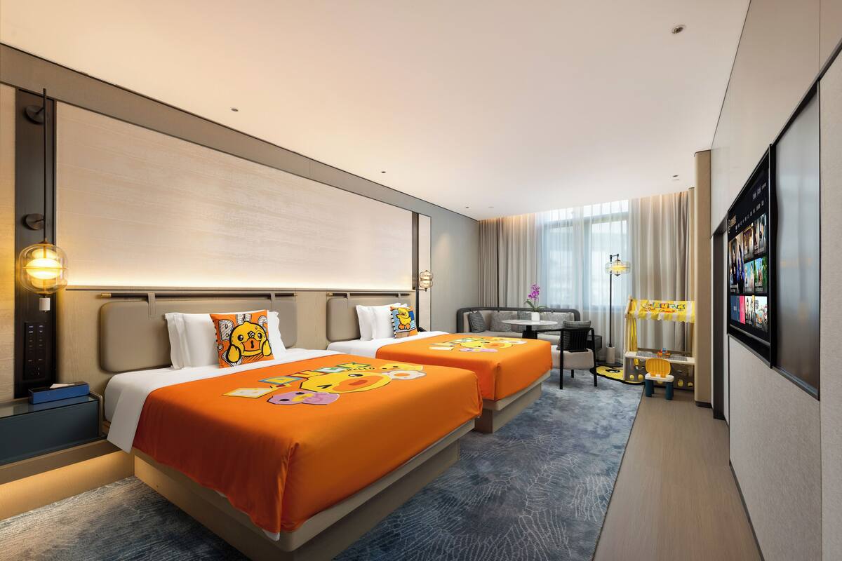 A hotel room with two beds, a desk, a TV, and a small play area.