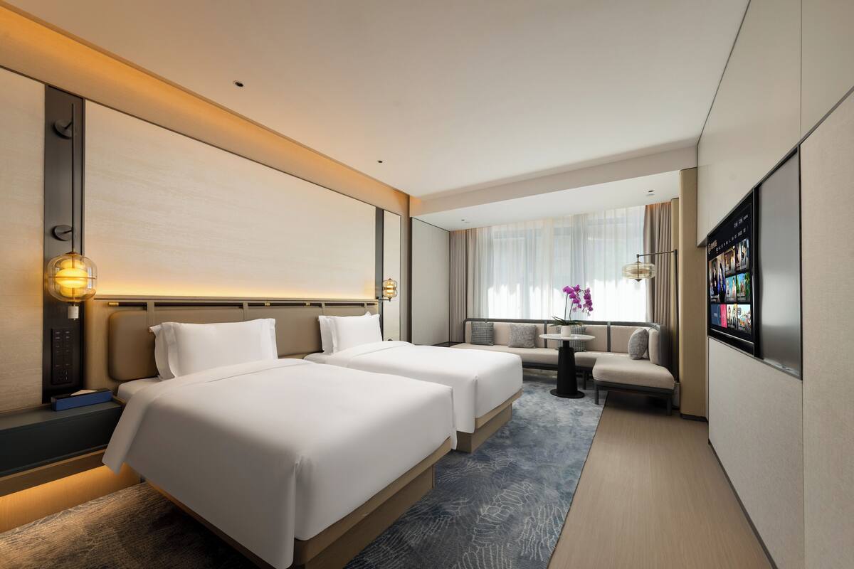 A hotel room with two beds, a sofa, a small table, a TV, and a window with curtains.