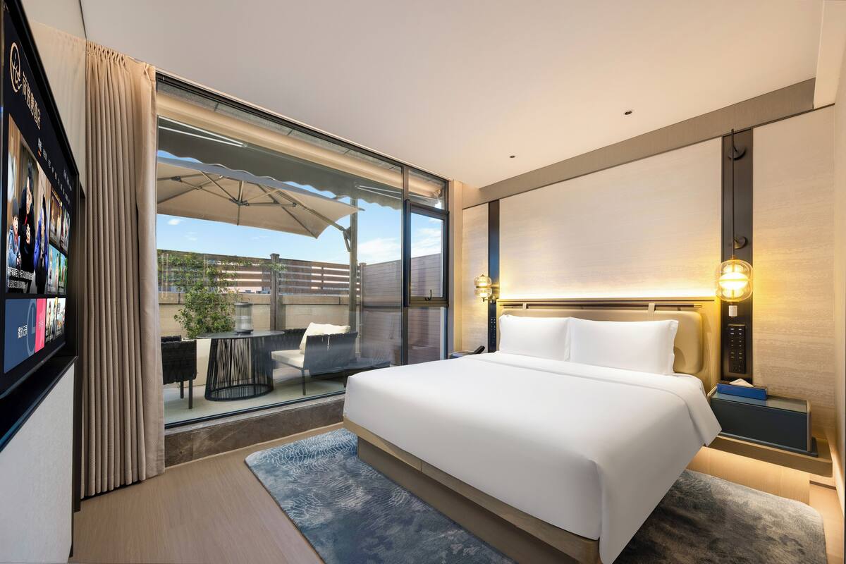 A modern hotel room with a large bed, a TV, a desk with a chair, and a view of the outdoors.