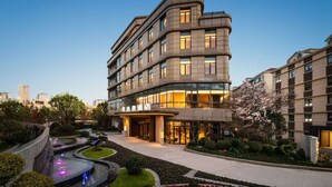 Un hôtel moderne à la façade incurvée, doté de grandes fenêtres et d’un jardin bien entretenu agrémenté d’un élément décoratif aquatique.