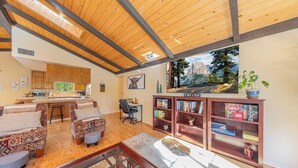 A spacious living area with a wooden ceiling, a kitchen in the background, and a bookshelf filled with books and games.