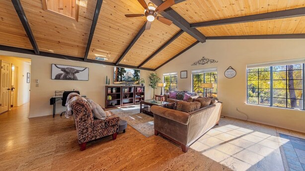 A spacious living room with wooden floors, a ceiling fan, and large windows offering a view of trees.