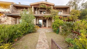 Une maison en pierre à deux étages, avec un chemin de pierre menant à l’entrée, le tout entouré d’une végétation luxuriante.
