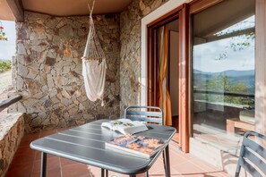 Ein Balkon mit einer Steinmauer, einem Hängesessel, einem Tisch mit einem Buch und Blick auf die Landschaft.
