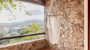Un balcon aux murs en pierre, agrémenté d’une décoration en macramé et offrant une vue sur un village.