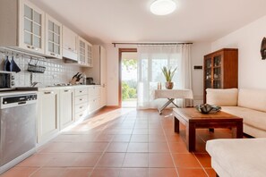 Una cocina moderna con armarios blancos, una zona de comedor con mesa y sofá, y vistas a través de un ventanal.