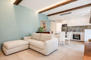 Un salon moderne avec un canapé d’angle, un coin repas avec une table et des chaises blanches, et une cuisine avec des meubles blancs et un dosseret carrelé.