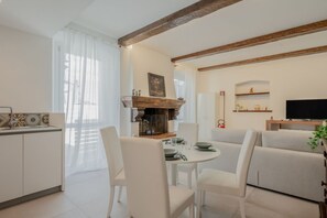A modern living room with a fireplace, a dining area with white chairs, a kitchen with a sink, and a TV on a wooden stand. - GuestHost - Mare Monti Apartment 1 (Dogana)