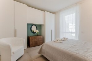 A bedroom with a bed, a white armchair, a wooden dresser, and a mirror. - GuestHost - Mare Monti Apartment 1 (Dogana)